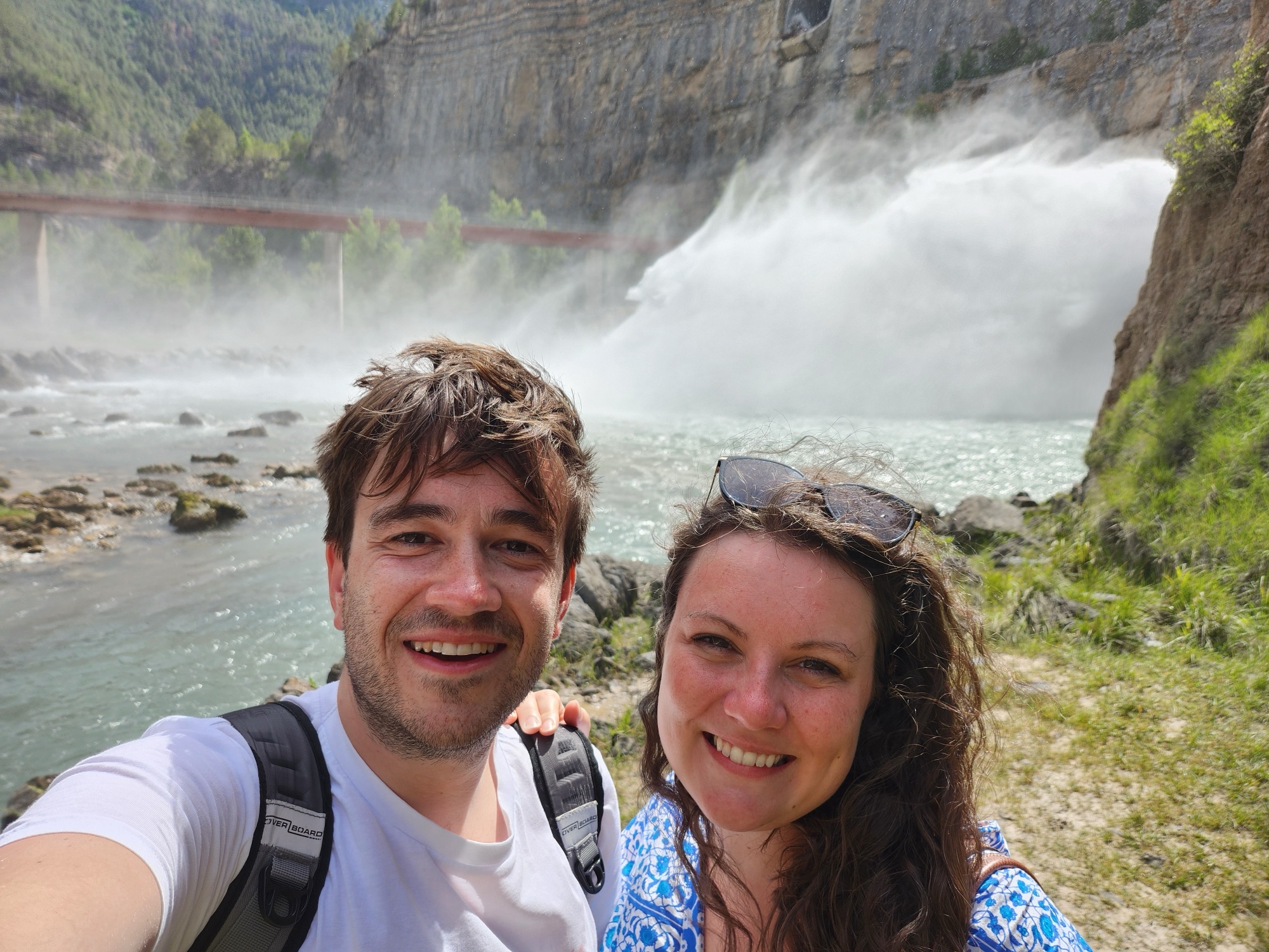 Beth and Harry smiling together beside a turquoise river, with a waterfall and canyon cliffs behind them.